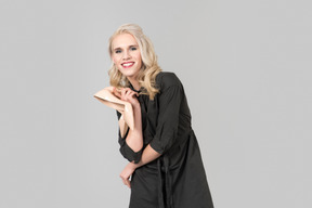 A young blond-haired person in a black dress and beige high heels in their hands standing against plain grey background