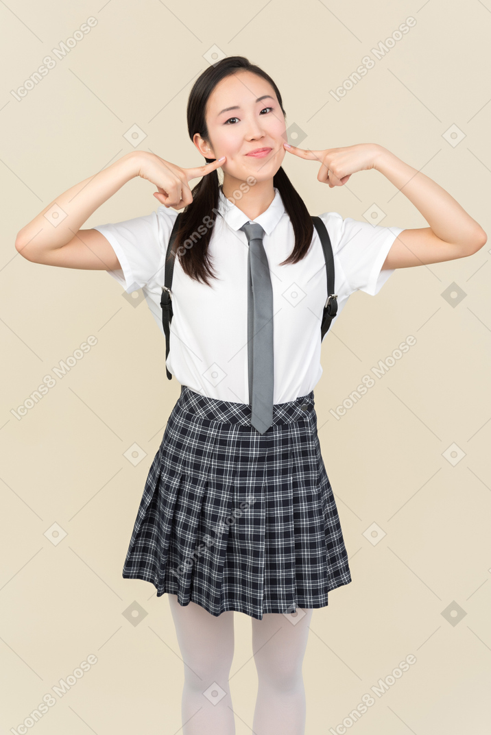 Asian school girl pointing at cheeks with fingers of both hands