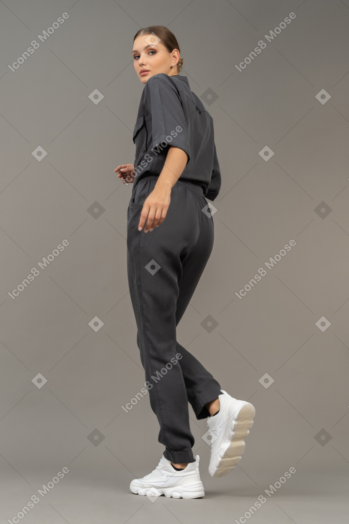 Three-quarter back view of a walking young woman in a jumpsuit looking at camera