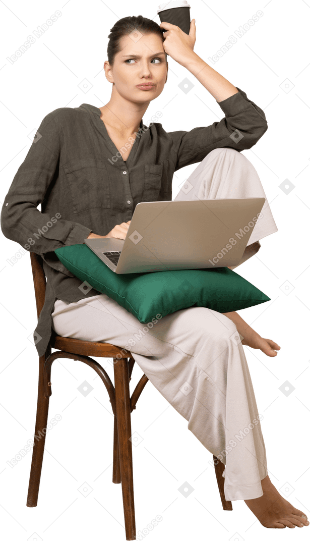 Vista frontal de una mujer joven confundida vistiendo ropa de casa sentado en una silla con una computadora portátil y café