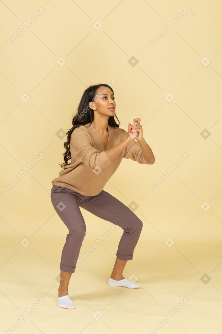 Вид в три четверти сидящей на корточках темнокожей молодой женщины с широко расставленными ногами