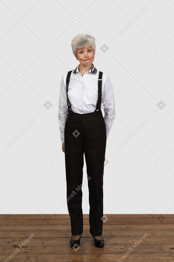 Vista frontal de una anciana perpleja en ropa de oficina haciendo muecas con las manos detrás de la espalda