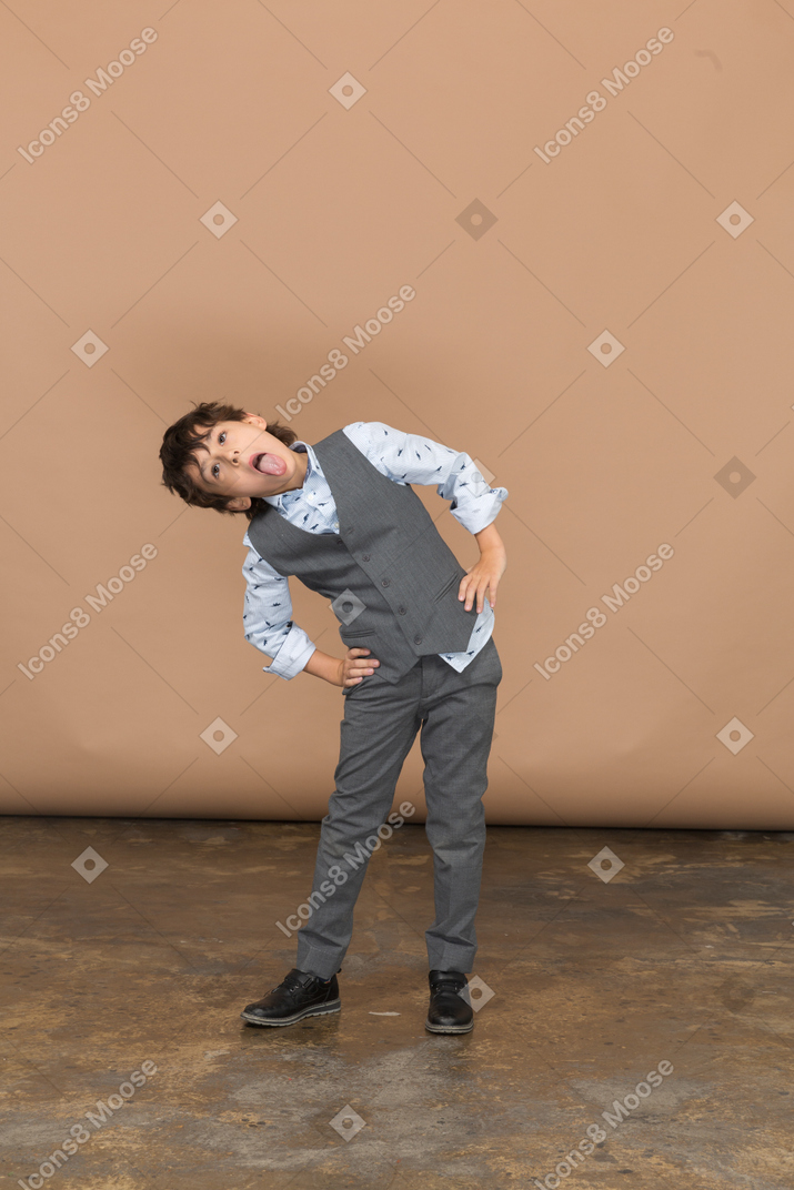 Vista frontal de un niño en traje posando con las manos en las caderas y mostrando la lengua
