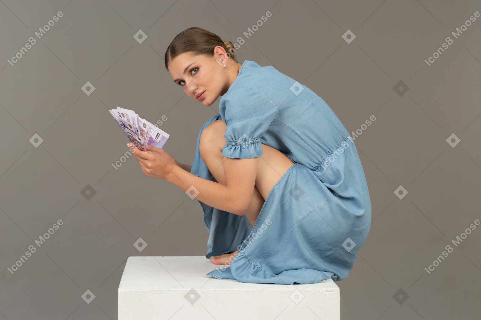 Side view of young woman sitting on cube with bunch of banknotes