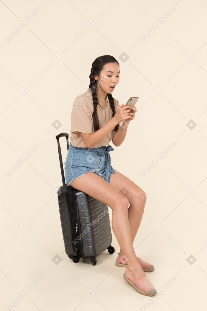 Surprised young female sitting on suitcase and using phone