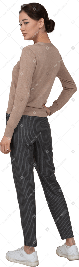 Vista posterior de tres cuartos de una mujer astuta en suéter y pantalones poniendo la mano en la cadera