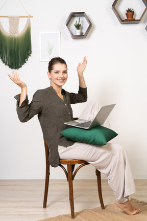 Vista de tres cuartos de una mujer joven vestida con ropa de casa sentada en una silla con una computadora portátil y levantando las manos