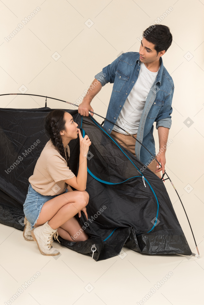 Interracial couple setting up a tent and woman showing a silence gesture