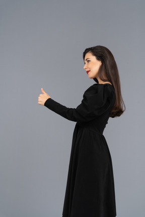 Vista lateral de uma jovem sorridente em um vestido preto com o polegar para cima