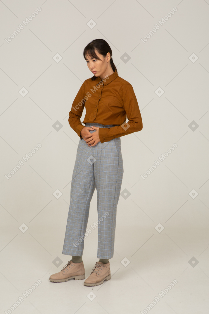Vista de três quartos de uma jovem mulher asiática de calça e blusa tocando a barriga