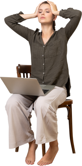 Dreiviertelansicht einer beschäftigten jungen frau mit kopfschmerzen, die mit einem laptop auf einem stuhl sitzt