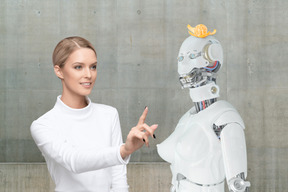 Blond woman wants to touch the robot with mandarine on the head
