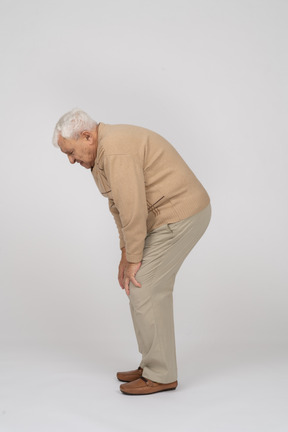 Side view of an old man in casual clothes bending down and touching his hurting knee