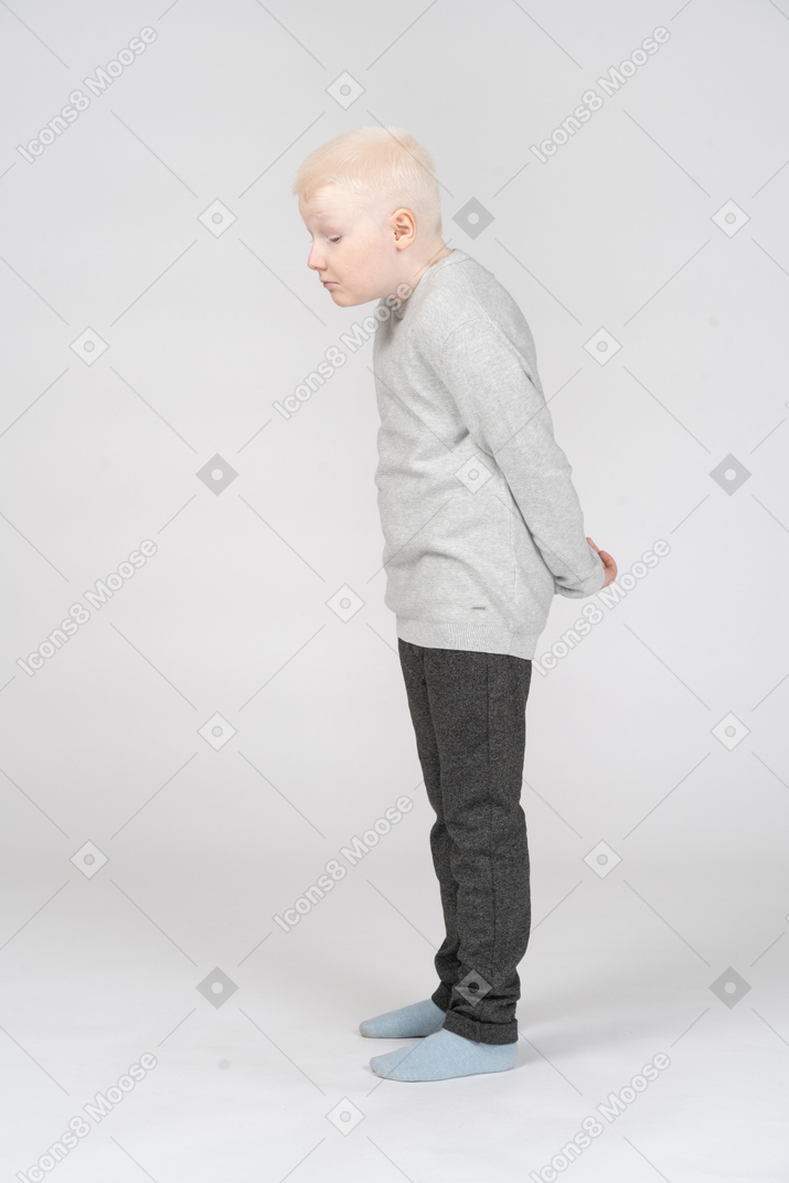 A blonde little boy in casual clothes standing and hiding hands behind