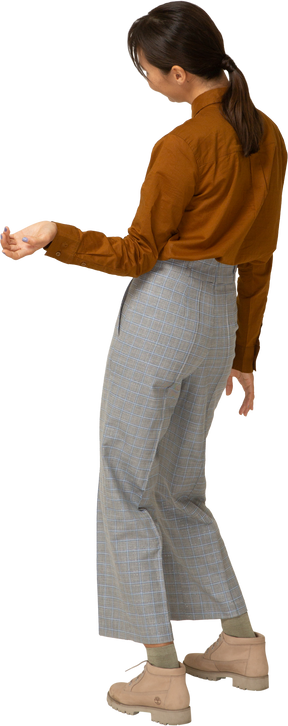 Vista posterior de tres cuartos de una joven mujer asiática en calzones y blusa inclinando la cabeza y extendiendo la mano