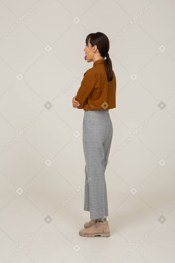 Vista traseira a três quartos de uma jovem mulher asiática de calça e blusa, cruzando os braços e mostrando a língua