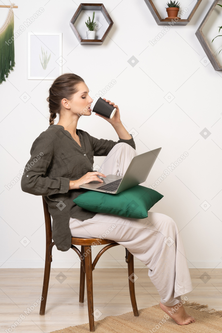 Side view of a perplexed young woman sitting on a chair and holding her laptop & drinking coffee