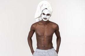 A young black man in grey trousers and a towel on his head going about his skin care routine