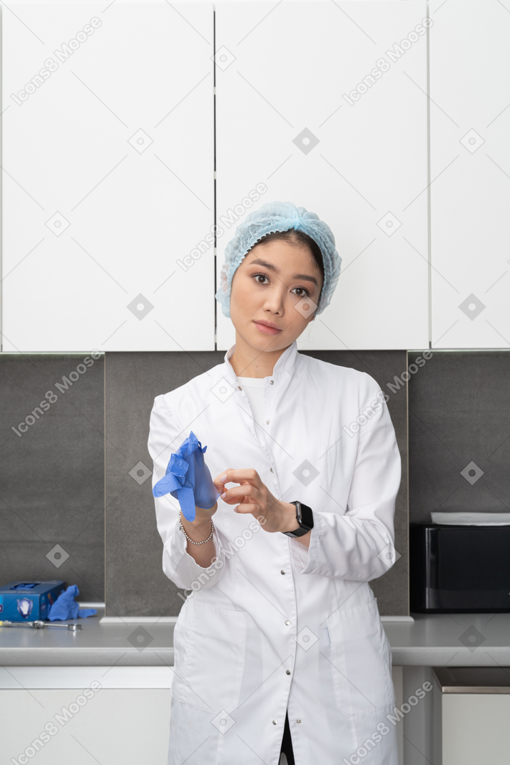 Front view of a young female doctor putting on protective gloves in her medical cabinet