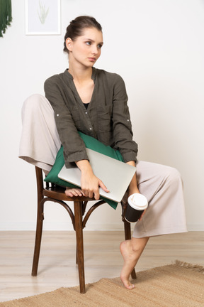 Vue de face d'une jeune femme assise sur une chaise et tenant son ordinateur portable et touchant une tasse de café