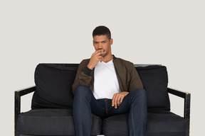 Front view of young man sitting on a sofa and holding cigarette in mouth