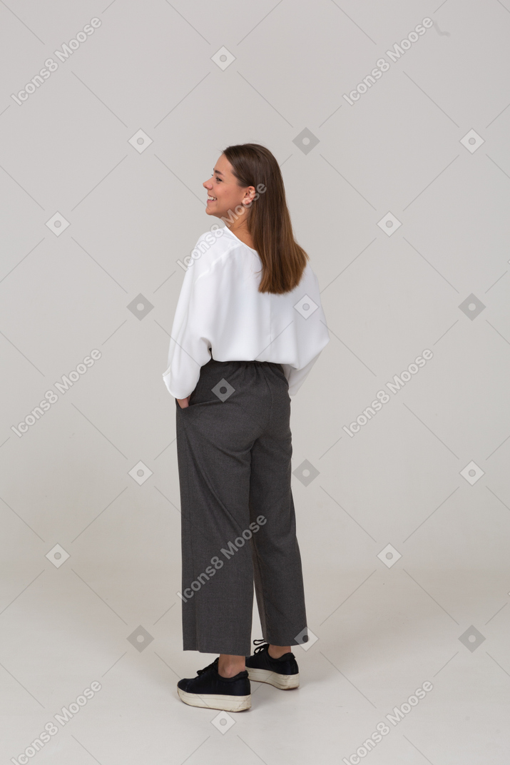Vista posterior de tres cuartos de una joven sonriente en ropa de oficina poniendo las manos en los bolsillos