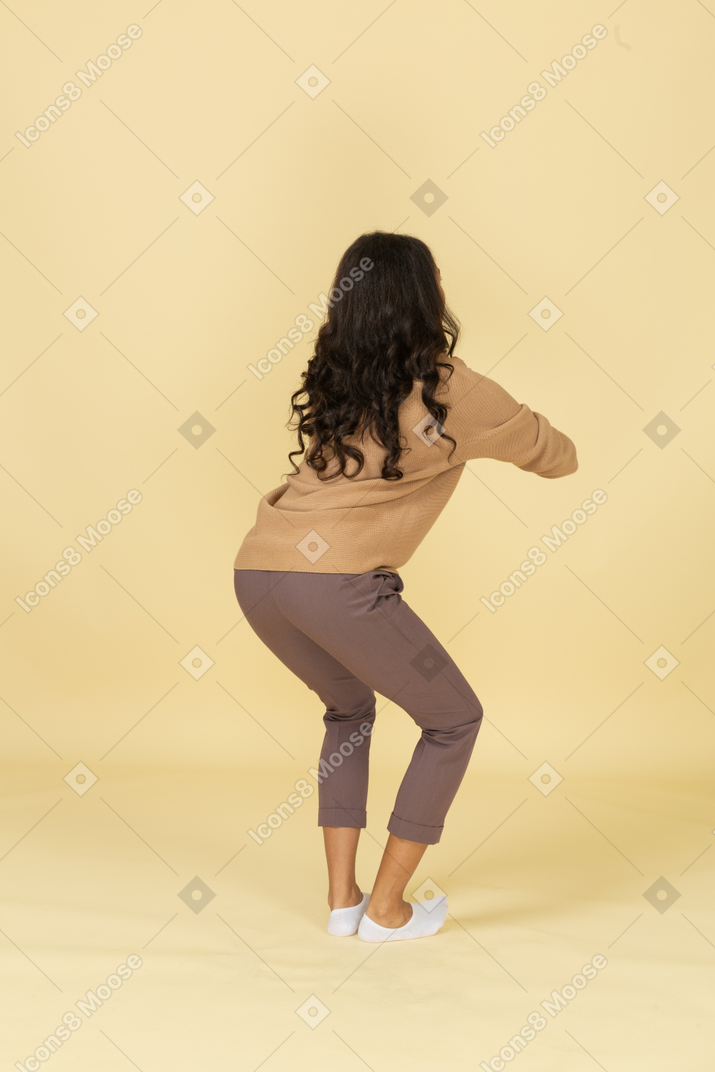 Three-quarter back view of a squatting dark-skinned young female holding hands together