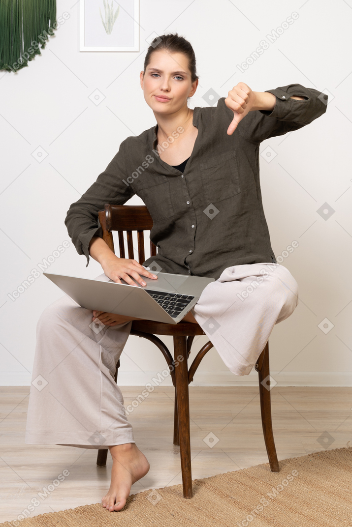 Вид спереди недовольной молодой женщины, сидящей на стуле с ноутбуком и показывающей большой палец вниз