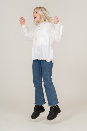 Вид в три четверти удивленной молодой женщины в повседневной одежде, прыгающей и поднимающей руки