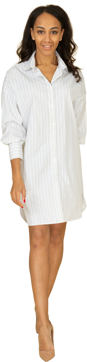 Vista frontal de una mujer joven de piel oscura caminando sonriente en vestido blanco