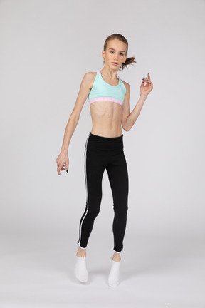 Vista frontal de uma adolescente em roupas esportivas, levantando a mão e olhando para a câmera enquanto pula