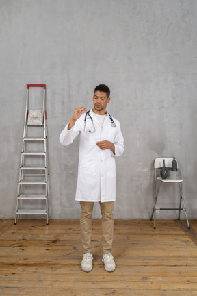 Front view of a young doctor standing in a room with ladder and chair showing a size of something