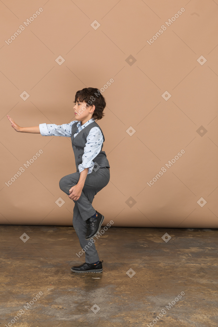 一个穿着灰色西装的男孩站在一条腿上并显示停止手势的侧视图