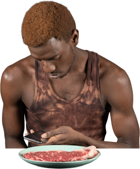Front view of a young afro man peeping in his phone before eating meat