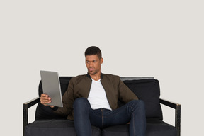 Vorderansicht eines gelangweilten jungen mannes, der auf einem sofa sitzt, während er das tablet beobachtet