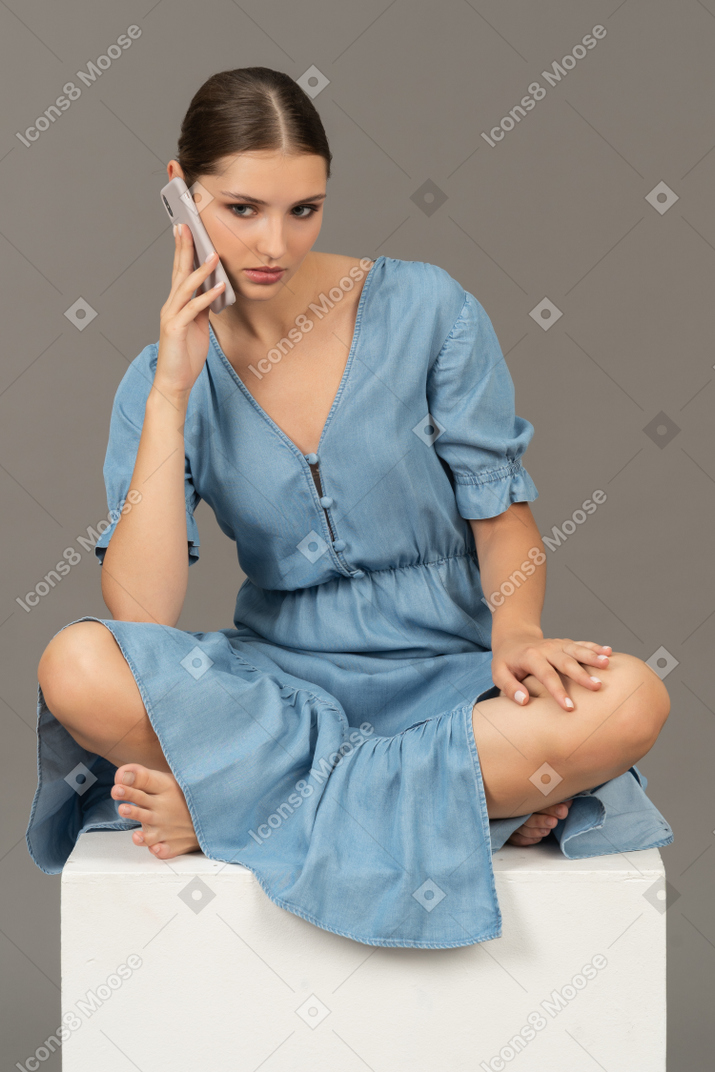 年轻女子坐在立方体上，用智能手机说话的正面图