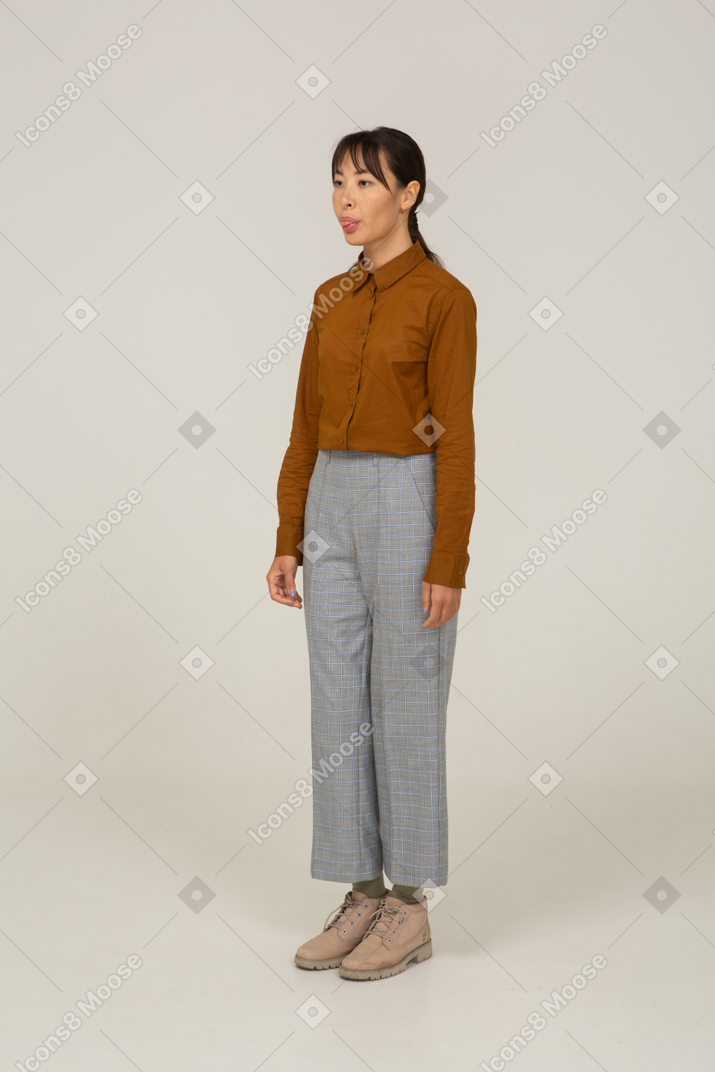 Vista di tre quarti di una giovane donna asiatica in calzoni e camicetta che mostra la lingua
