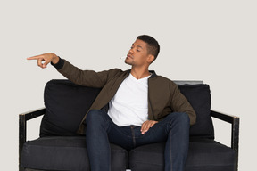 Vista frontal de un joven arrogante sentado en un sofá y sosteniendo el cigarrillo en la boca