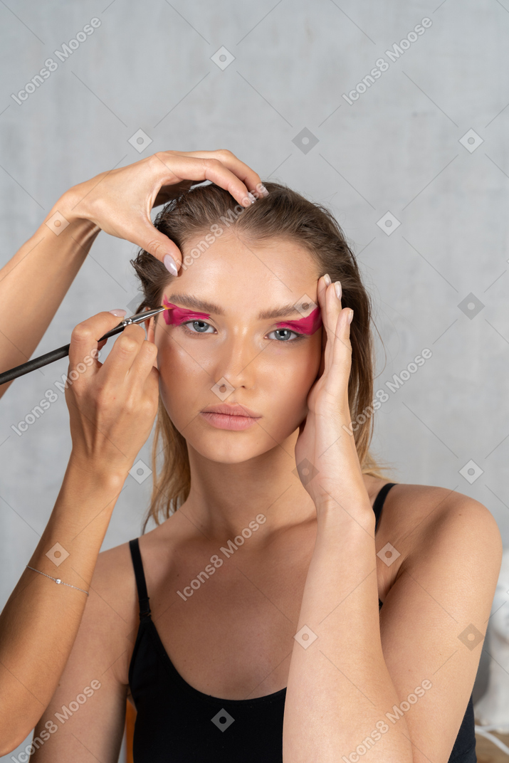 Front view of a young woman touching temple & make-up artist applying eyeshadow