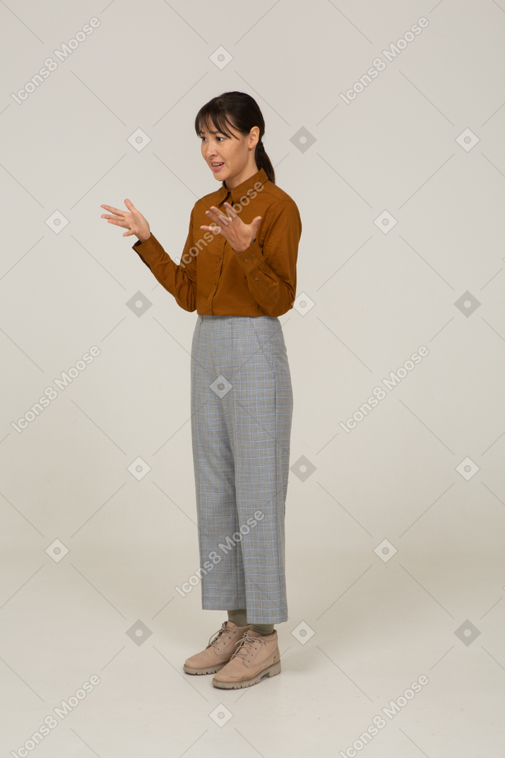 Vista di tre quarti di una giovane donna asiatica interrogativa in calzoni e camicetta che alza le mani