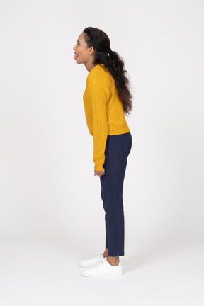 Garota com roupas casuais em pé em seu perfil e mostrando a língua