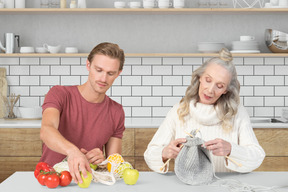 A man taking out fruits from a bag next to a knitting woman in the kitchen