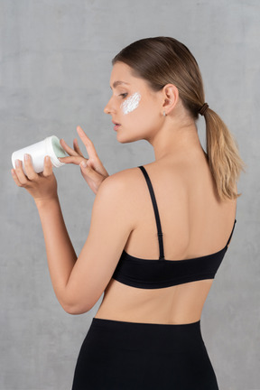 Back view of a young woman applying face cream on her cheekbones