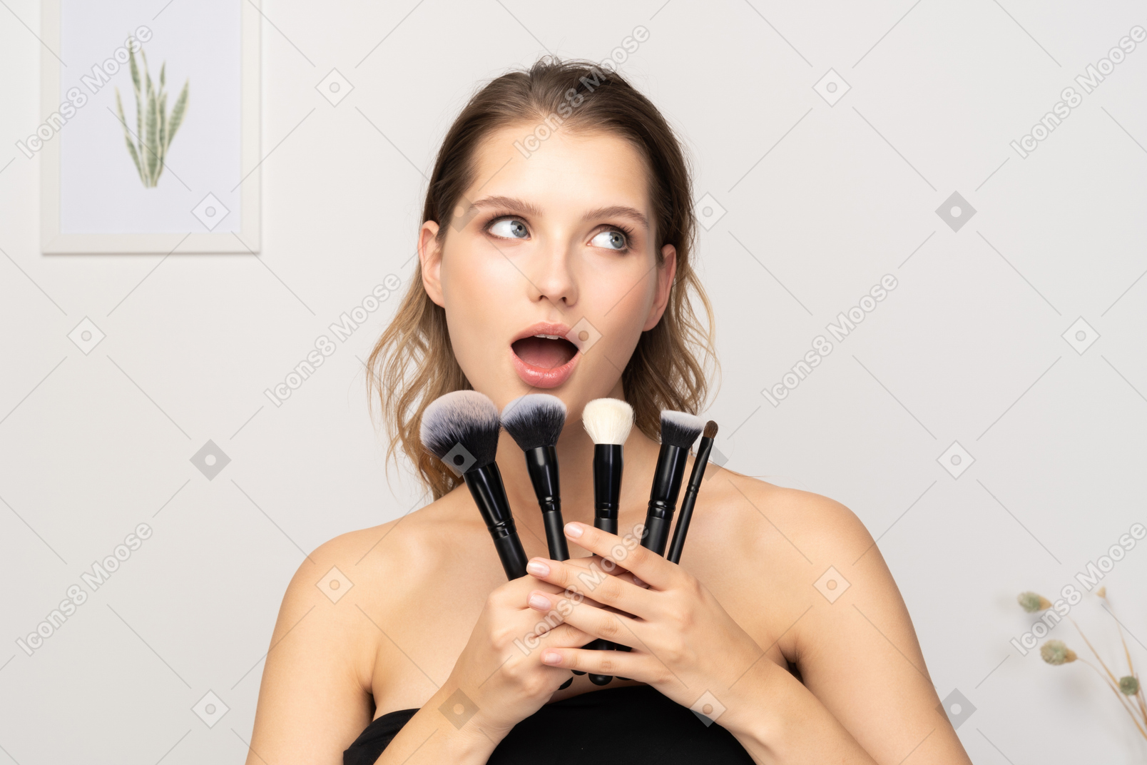 Vista frontal de una mujer joven sorprendida sosteniendo pinceles de maquillaje