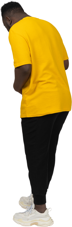 Vista posteriore di tre quarti di un giovane uomo dalla pelle scura in maglietta gialla che tocca lo stomaco e guarda in basso