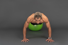 Vista frontal de un hombre afro sin camisa haciendo flexiones en una pelota de gimnasia