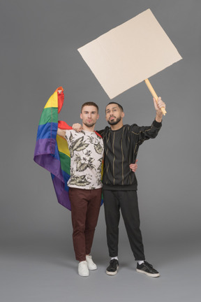 Vista frontal de dois jovens meio abraçando e levantando um outdoor e uma bandeira lgbt