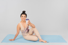 Молодая индийская гимнастка сидит на циновке йоги и держит чашку кофе