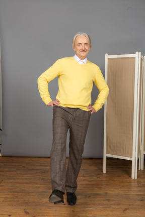 Front view of a smiling old man putting hand on hips while raising leg