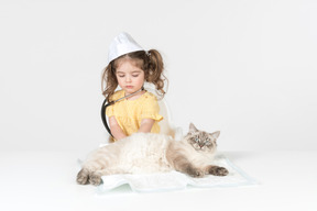 Little kid girl with stethoscope and wearing medical hat curing a cat